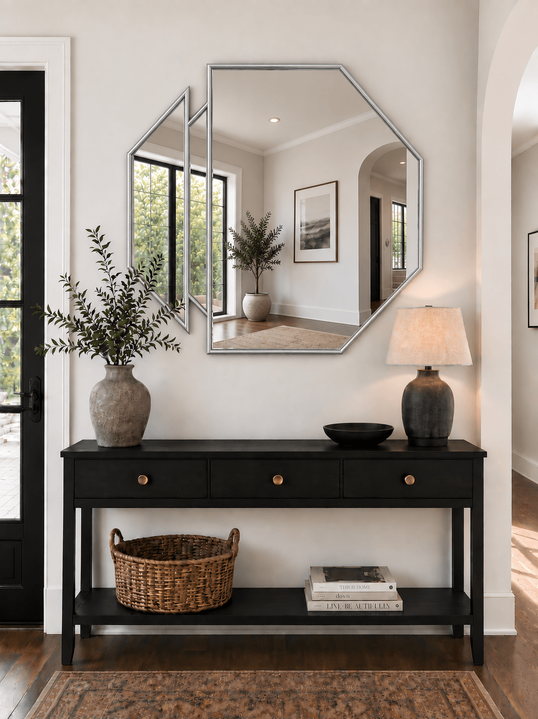 An octagonal wall mirror with a silver frame hangs above a black console table in a bright entryway. The mirror reflects the interior hallway, while the table is styled with a vase of greenery, a lamp, a bowl, a woven basket, and books.