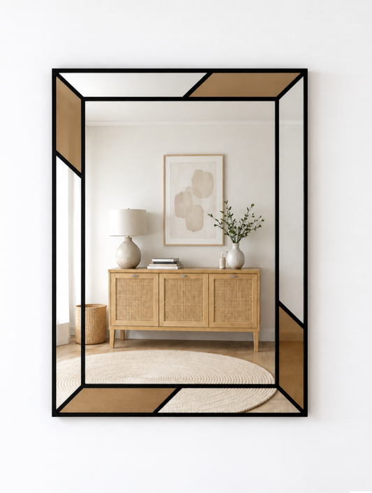 A rectangular wall mirror with a black metal frame and geometric tan inset panels, shown hanging on a white wall. The mirror reflects a styled interior scene with a light wood cabinet, lamp, framed art, and a rug.