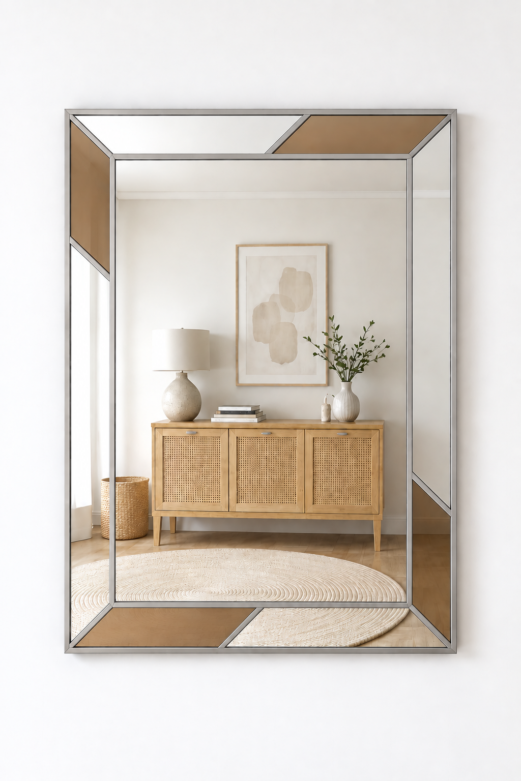 A large rectangular wall mirror with a geometric frame, featuring alternating clear and warm taupe-beige glass panels surrounded by a brushed silver metal border. The mirror is mounted on a clean white wall and reflects a beautifully styled interior room with a light wood credenza, a white table lamp, and a round beige rug.
