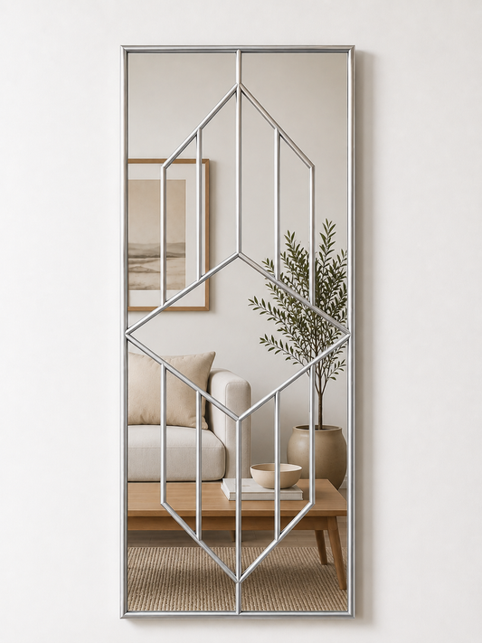 A tall rectangular wall mirror with a thin silver geometric frame overlay, shown hanging on a light beige wall. The mirror reflects a styled living room interior with a sofa, coffee table, plant, and framed art.