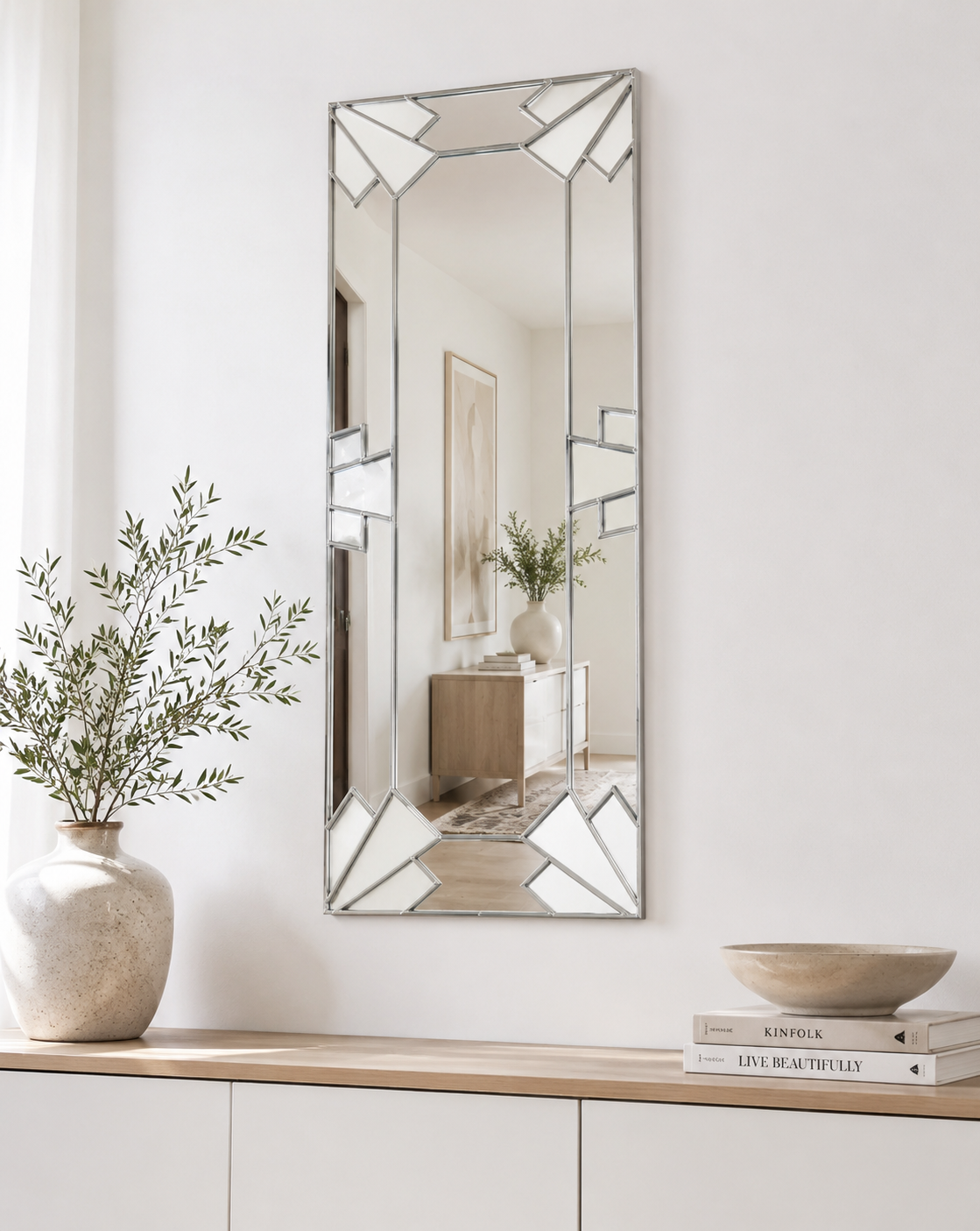 A tall rectangular wall mirror with a geometric silver frame hangs on a white wall above a light wood console table. The mirror reflects a softly lit interior, and the scene is styled with a ceramic vase of greenery on the left and stacked books with a bowl on the right.