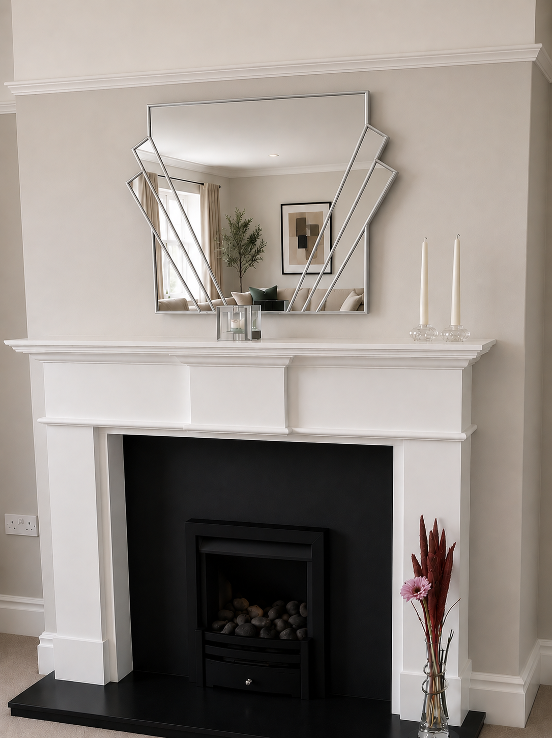 A white fireplace mantel with a black firebox insert, a black hearth, and a large geometric silver-framed mirror mounted above. The room has beige walls, and decorative items including two white taper candles and a vase with dried pink flowers sit on the mantel and hearth.