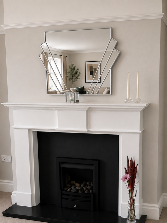 A white fireplace mantel with a black firebox insert, a black hearth, and a large geometric silver-framed mirror mounted above. The room has beige walls, and decorative items including two white taper candles and a vase with dried pink flowers sit on the mantel and hearth.