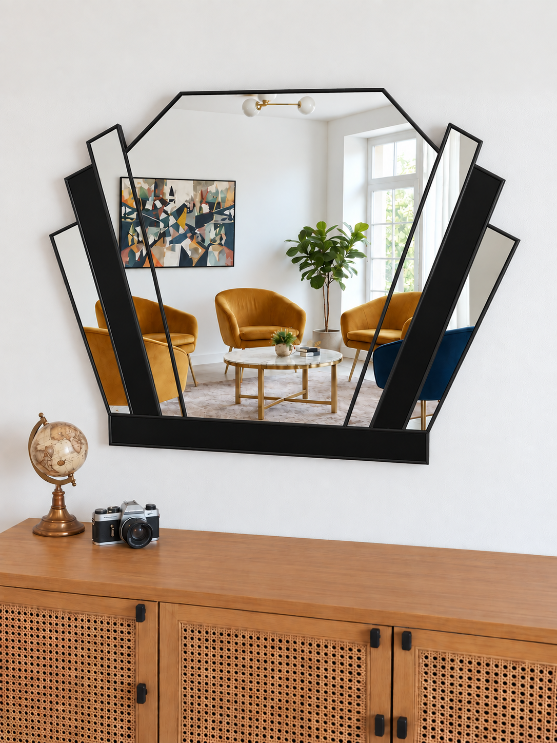 A large black-framed geometric wall mirror with multiple angled panels, mounted on a white wall above a wooden cabinet. The mirror reflects a modern living room with mustard yellow armchairs, a coffee table, and a green plant.