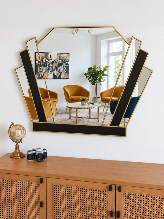 A large geometric wall mirror with a gold frame and black inner panels, mounted on a white wall above a wooden cabinet. The mirror reflects a modern living room with mustard yellow armchairs, a white coffee table, and a green plant.