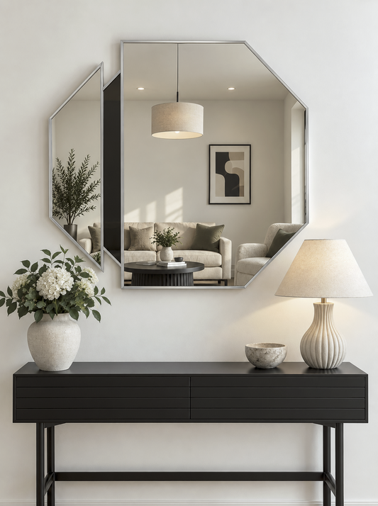 An octagonal wall mirror with a silver frame hangs above a black console table in a bright, modern room. The mirror reflects a living room background, and the table is styled with a white vase of flowers, a lit table lamp, and a small bowl.