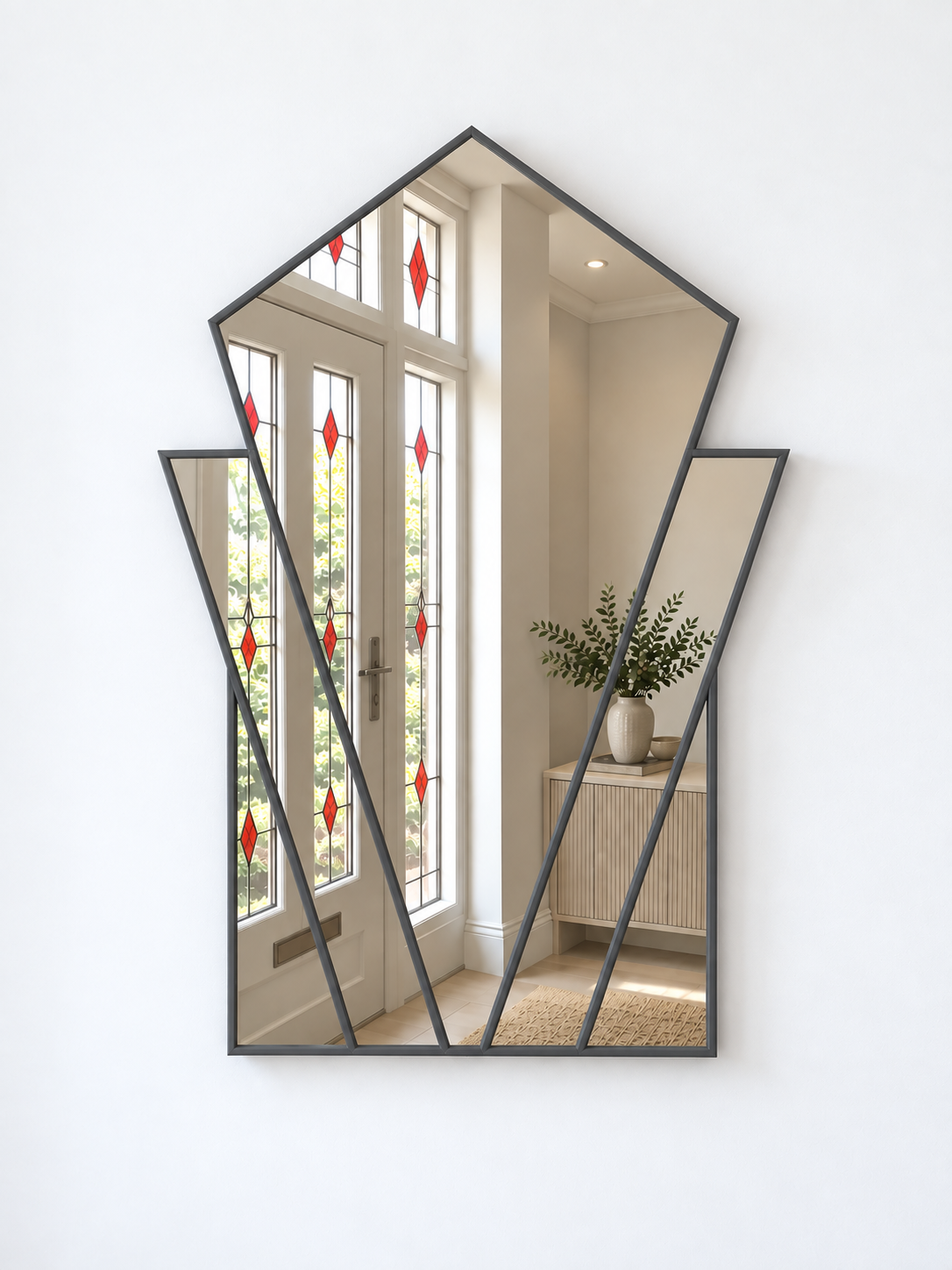 A modern geometric wall mirror with a dark charcoal gray frame, mounted on a clean white wall. The mirror features multiple angled panels reflecting a bright hallway with white doors and a wooden cabinet.