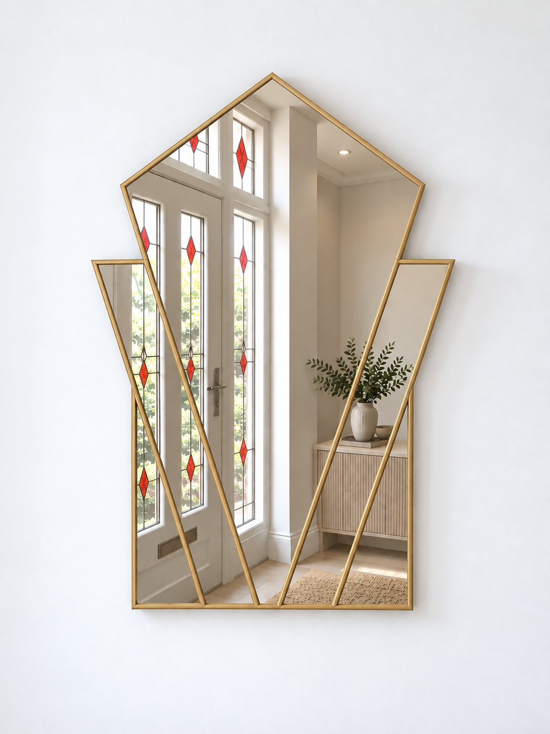 A geometric gold-framed wall mirror with multiple angled panels, mounted on a white wall. The mirror reflects a bright interior hallway with white doors, red diamond-shaped stained glass accents, and a light wood cabinet with a potted plant.