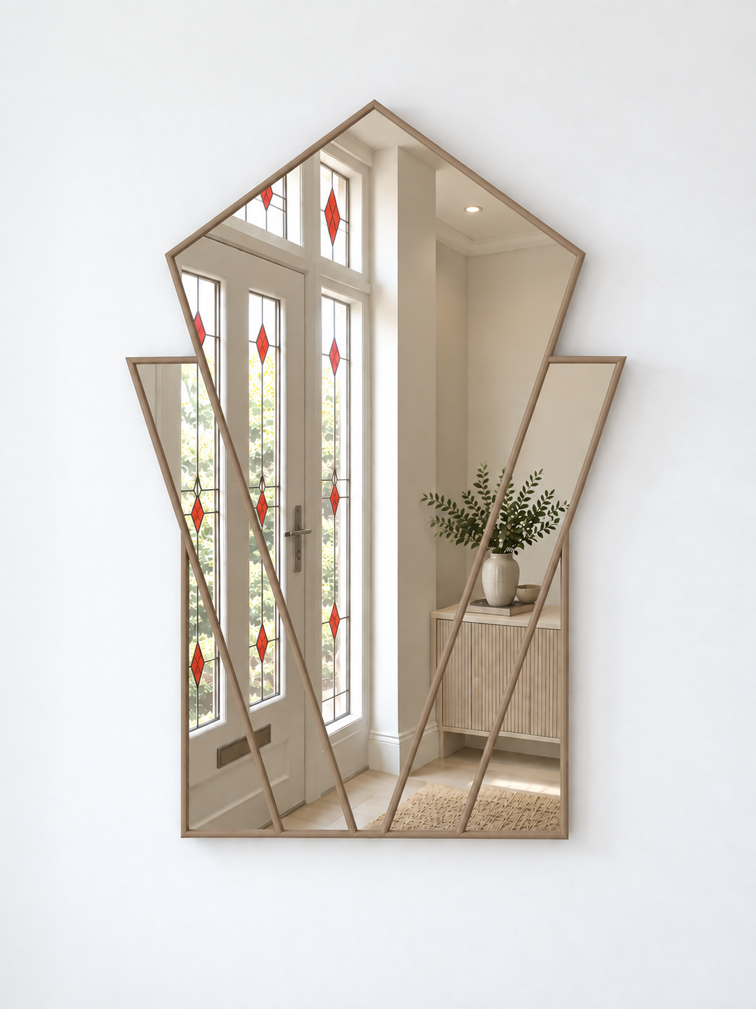 A modern geometric wall mirror with a light natural wood frame, mounted on a white wall. The mirror is shaped like a stylized house, reflecting a bright entryway with white double doors featuring red diamond-shaped stained glass panels and a wooden sideboard with a potted plant.