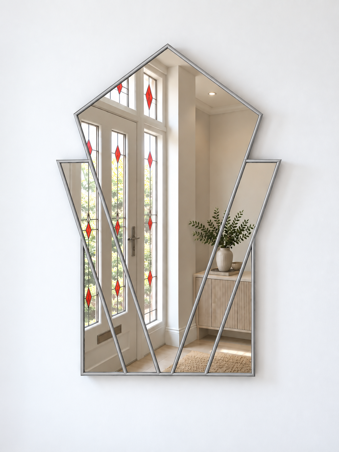 A modern geometric wall mirror with a silver frame, mounted on a white wall. The mirror is shaped like a stylized house and reflects a bright interior hallway with white doors featuring red diamond-shaped stained glass panels.