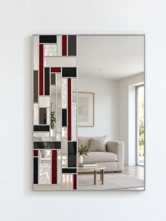 A rectangular wall mirror with a silver frame and an abstract geometric pattern of black, red, grey, and textured glass panels on the left side